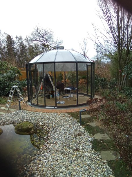 Glassund Metallgartenhaus umgeben von einem Garten mit Steg und Steinen.